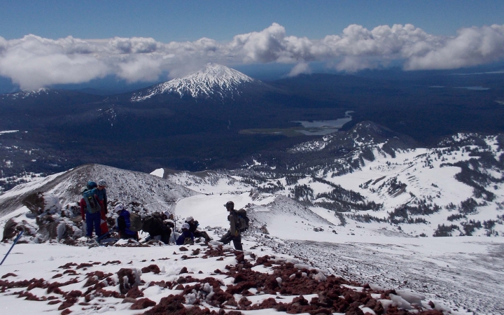 A person wearing a backpack stands on a snowy and rocky outlook, looking out over a vast mountainous landscape.